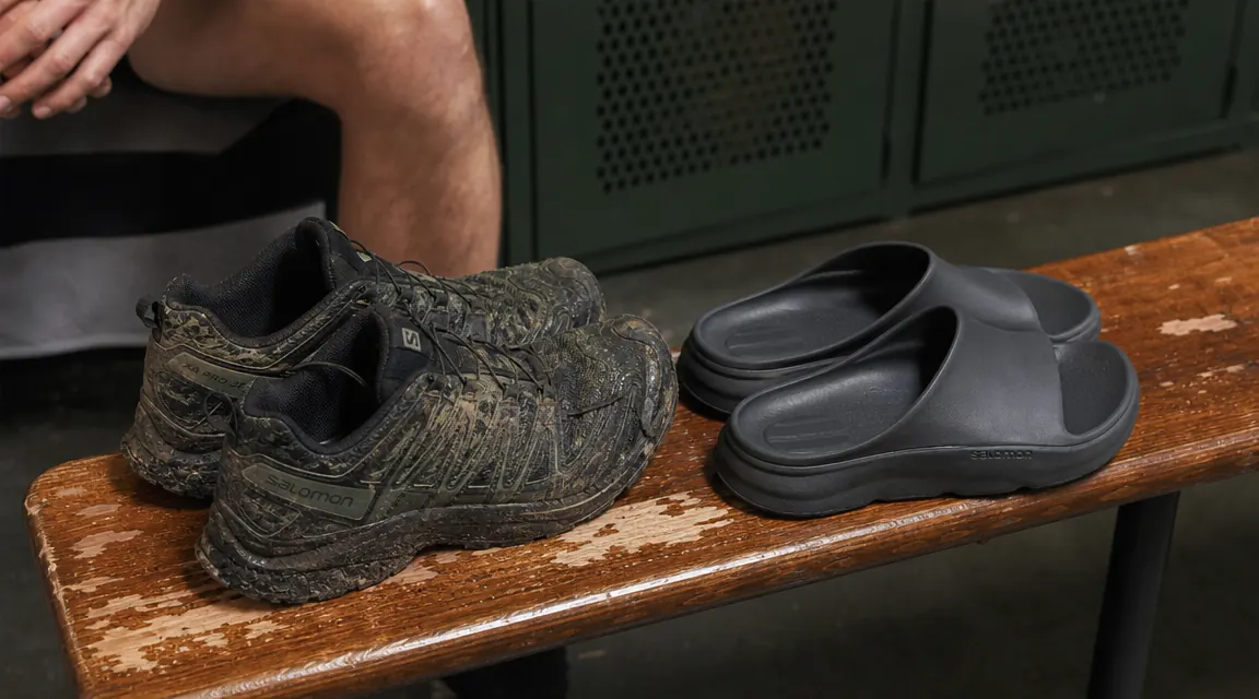 Pair of OOFOS OOriginal recovery sandals sitting on a gym bag next to trail running shoes, side by side on a gym locker room bench